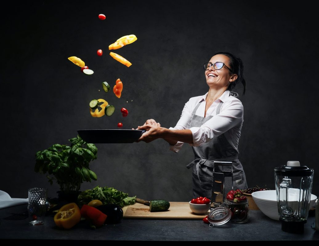 Happy middle-aged female chef tossing chopped vegetables in the air from a frying pan